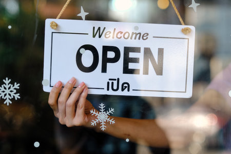 A hand holds a bilingual Welcome Open sign on a glass door with snowflake decals, indicating store...