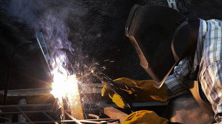 Welder wearing helmet and gloves focuses on fusing metal with bright arc light, flying sparks, and...