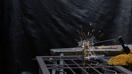 Worker in gloves performs welding on a metal frame, creating visible sparks and smoke during...