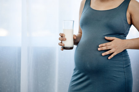 Close-up of a pregnant woman in a gray dress holding her belly and a glass of milk, focusing on prenatal health and careの写真素材
