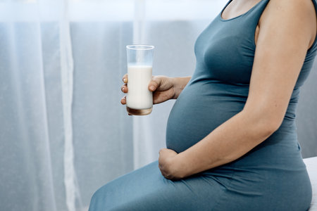 Close-up of a pregnant woman in a blue dress holding her belly and a glass of milk, symbolizing prenatal nutrition and careの写真素材
