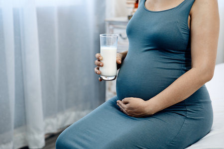 Close-up of a pregnant woman in a blue dress sitting indoors, holding her belly with one hand and a glass of milk in the otherの写真素材