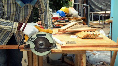 Worker in protective gloves and mask uses an electric circular saw to cut a wooden plank on outdoor...