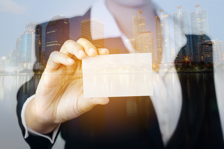 Double exposure of business woman hand showing a blank empty name card on city buildings background.の写真素材