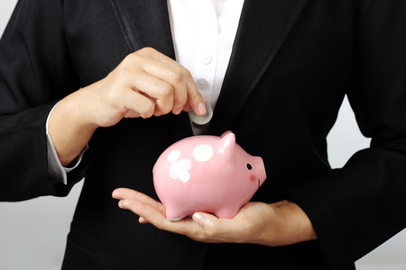 Close up of businesswoman putting a coin into piggy bank on gray background, finance theme.の写真素材