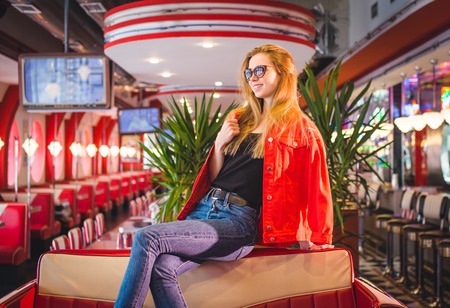 young stylish beautiful woman sitting in city cafe in red coat, street style, autumn trend, warm...