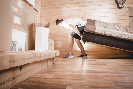It's moving day! Busy funny guy in casual clothes is carrying big boxes in his hands and trying to keep one using his head, isolated on yellow backgroundの写真素材