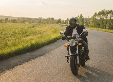 Bearded biker in sunglasses, helmet and black leather clothing riding cruiser motorcycle along asphalt road winding among tall green trees on sunny summer evening.の写真素材