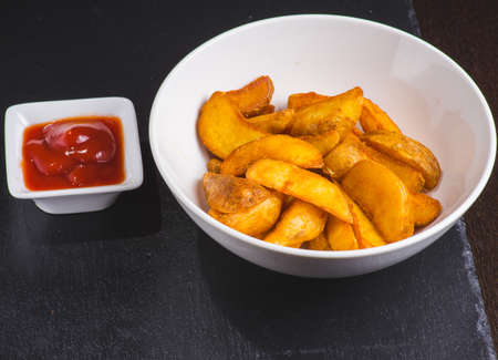 Rustic potatoes with ketchup on light wooden plate on dark wooden background.の写真素材