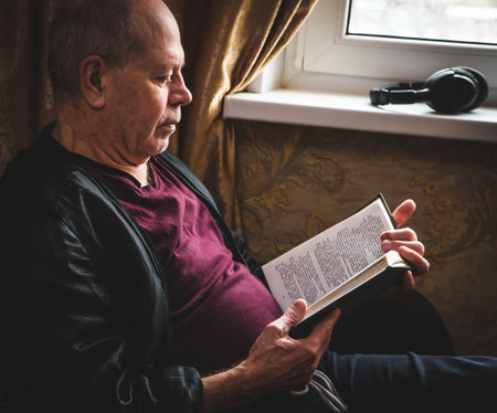 Profile side view of nice focused sad tired bored stylish old man near window wearing checked shirt reading modern book in white light modern interiorの写真素材