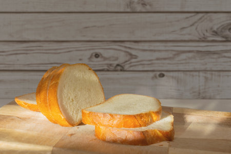 Freshly baked loaf of bread sliced into thick pieces on a wooden cutting board in a cozy kitchen setting under the morning sunshine.の写真素材
