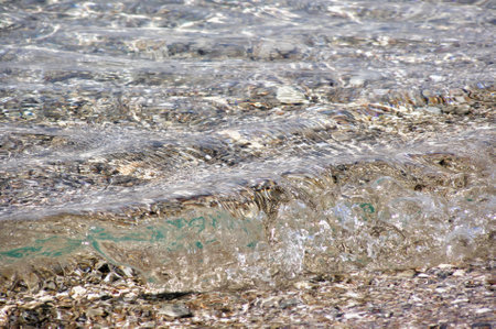 Crystal clear water ripping on rocky shoreline. Transparent water waves gently lapping over a bed of pebbles and seashells at the edge of a serene rocky beach.の写真素材