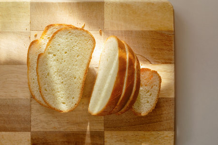 Morning light bathes a wooden cutting board as slices of fresh bread lie invitingly. The soft glow enhances the rustic charm, creating a warm culinary atmosphere.の写真素材