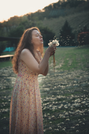 Beautiful young girl playing in dandelion field in beautiful spring warm sunset. Wallpaper full of joy and happinessの写真素材