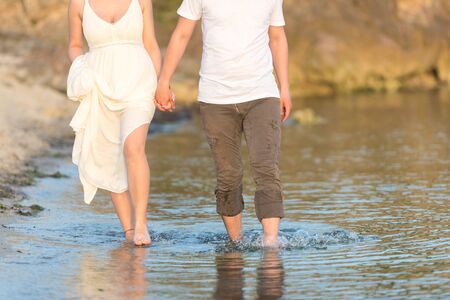 Outdoor shot of romantic young couple walking along the sea shore holding hands. Young man and woman walking on the beach together at sunset, body closeupの写真素材