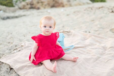 Cute baby girl in a red dress sitting and playing on the beach with sandの写真素材