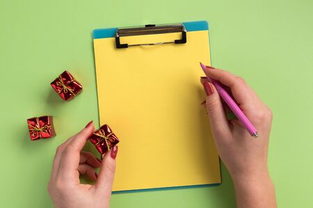 Christmas or New Year planning concept. Clipboard, pen, small gifts and female hands, on green...