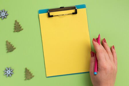 Female hand holding pen, clipboard with yellow sheet and christmas decor, on green background. Flat lay, top view, copy space.の写真素材