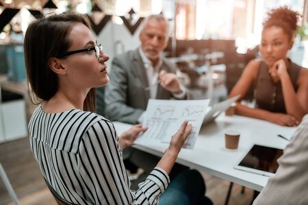 Analyzing results. Young business woman holding financial report and discussing something with colleagues while sitting at the office table together. Brainstorming. Teamworkの写真素材