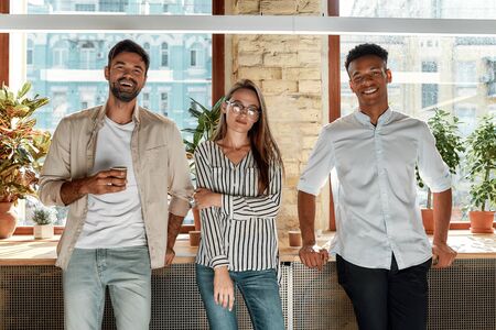 Nice coffee! Three cheerful colleagues in casual wear looking at camera while standing in the modern office. Working together. Teamの写真素材