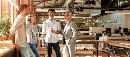 Project done. Business people holding coffee cups and talking about something with smile while standing in the modern officeの写真素材