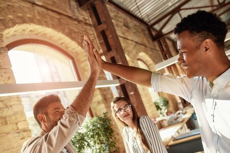 Celebrating success. Two cheerful men giving high-five while standing with colleagues in the modern officeの写真素材