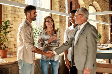 Nice to meet you! Two business people shaking hands and smiling while standing with coworkers in the creative office.の写真素材