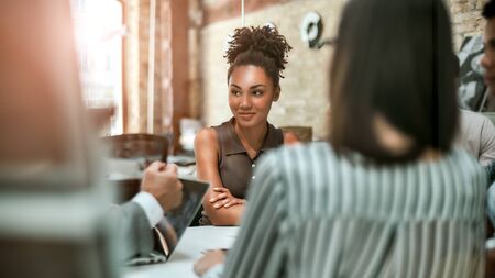Happy to work here. Young and cheerful afro american woman smiling and looking aside while having a meeting with colleagues in the modern officeの写真素材