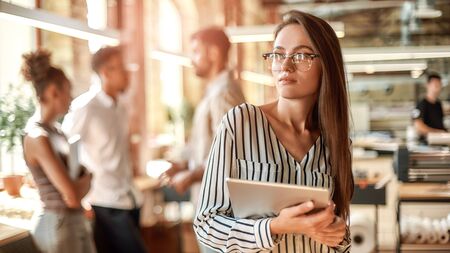 Smart and confident. Young woman in eyeglasses holding digital tablet and looking at camera while standing in the modern office. Teamwork. Office lifeの写真素材