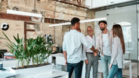 Welcome! Two men shaking hands and smiling while standing with coworkers in the creative office. Greeting. Teamworkの写真素材