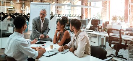 Working process. Mature businessman talking about something with his young colleagues while sitting together at the office table in the modern office. Teamwork. Business conceptの写真素材