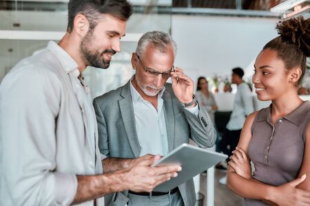 Let me see. Confident mature business man in formal wear looking at digital tablet while standing with young coworkers in the modern office. Work concept. Business conceptの写真素材