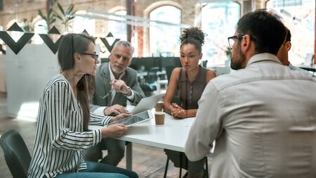 Using digital technologies. Young business woman holding digital tablet and discussing something with colleagues while sitting at the office table togetherの写真素材