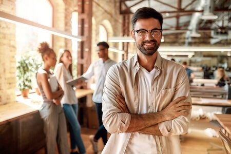 Great working day! Young bearded man in casual clothes keeping arms crossed and looking at camera with smile while standing in the creative officeの写真素材