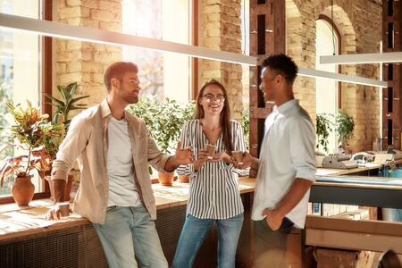 Nice coffee break. Three young and cheerful colleagues in casual wear holding coffee cups and discussing something while standing in the modern officeの写真素材
