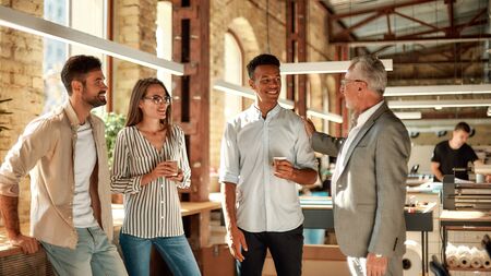 Good job! Smiling mature man in formal wear patting his young afro american colleague on shoulder while standing with coworkers in the modern officeの写真素材