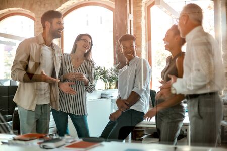 Happy at work. Group of cheerful people smiling and discussing something while standing in the modern office. Teamworkの写真素材