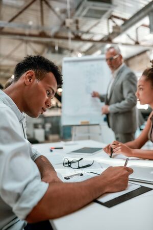 Important information. Young afro american man is writing something while his colleague explaining new business strategy with white board on the backgroundの写真素材