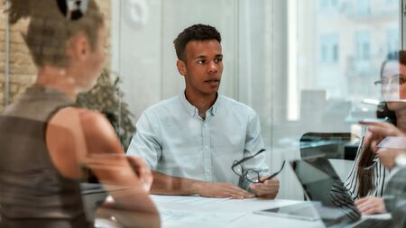 Business expert. Young afro american man holding eyeglasses and explaining something to his colleagues while sitting at the office table behind the glass wall in the modern officeの写真素材