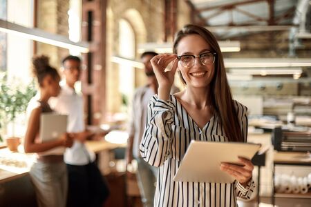 Successful business woman. Young cheerful woman holding digital tablet and looking at camera with smile while standing in the modern officeの写真素材