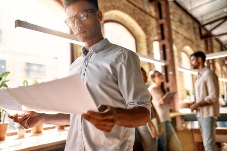 Working on new project. Young afro american man in casual clothes analyzing documents while standing in the modern officeの写真素材