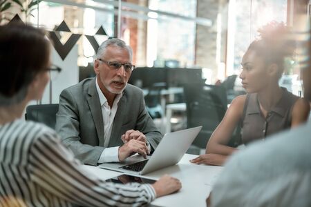 Brainstorming. Serious mature man in formal wear discussing something with his multicultural team...