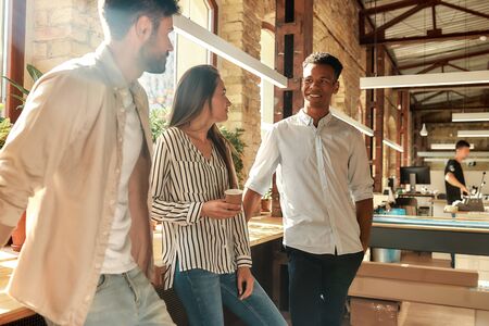Pause at work. Young and cheerful colleagues in casual wear talking about something and holding coffee cups while standing in the modern office. Coffee breakの写真素材