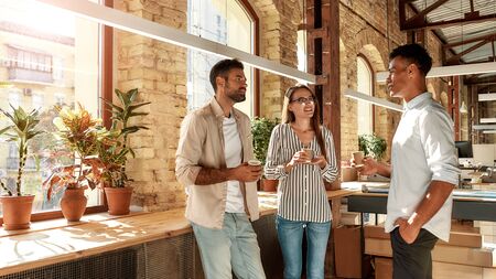 Nice talk. Three young colleagues in casual wear holding coffee cups and discussing something while standing in the modern office. Coffee break.の写真素材
