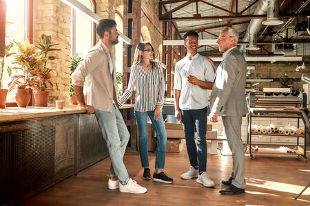 Well done! Cheerful mature man in formal wear patting his young afro american colleague on shoulder while standing with coworkers in the modern office. Teamwork. Office lifeの写真素材