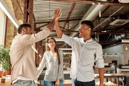 We did it! Two cheerful men giving high-five while standing with colleagues in the modern office. Teamwork. Office lifeの写真素材