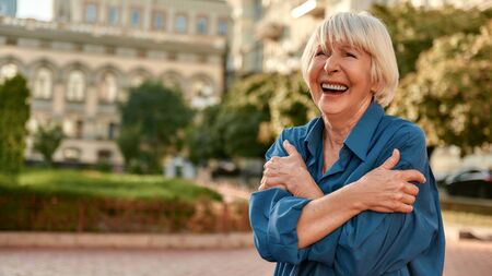 I am optimist. Beautiful senior woman in casual wear hugging her shoulders and smiling while standing outdoors on a sunny day. Old age. Happinessの写真素材