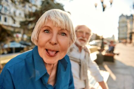 Feeling playful. Funny senior woman in casual clothes showing her tongue while sitting with husband on the bench. Positive emotionsの写真素材