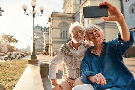 Happy moments. Cheerful senior couple in casual clothes making a selfie while sitting on the bench together. Modern technologies. Photo conceptの写真素材