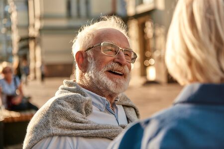 Happy senior bearded man in glasses looking at his wife and smiling while sitting on the bench together. Walking outdoors. Old ageの写真素材